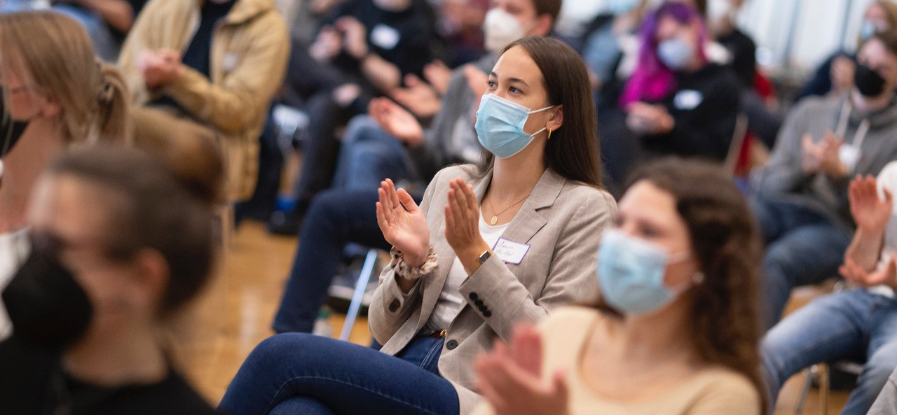 Das Bild zeigt eine Gruppe sitzender Menschen, die sich in einem großen Rum befinden und applaudieren. Die Personen im Vordergrund tragen blaue Atemschutzmasken. Der Hintergrund verschwimmt.