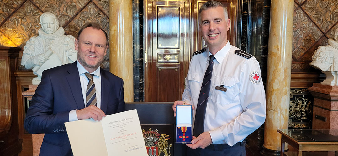 Das Bild zeigt zwei Personen in einem festlichen Raum. Die Person auf der rechten Seite trägt eine Uniform und nimmt eine Medaille entgegen. Die Person auf der rechten Seite trägt einen Anzug und hält eine Urkunde in der Hand und zeigt diese in die Kamera. Im Hintergrund ist eine weiße Büste zu sehen.