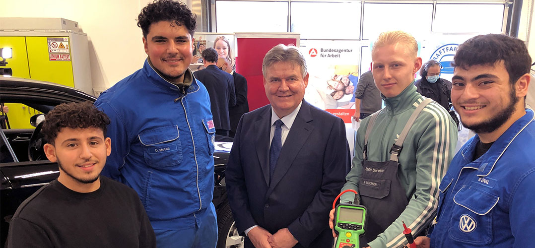 Das Bild zeigt fünf Personen im Vordergrund. Die Person in der Mitte trägt einen Anzug. Die Personen an den Rändern tragen Arbeitskleidung. Der Raum scheint eine Werkstatt zu sein und im Hintergrund sind Teile von einem Auto zu sehen sowie ein Banner mit der Aufschrift "Bundesagentur für Arbeit".