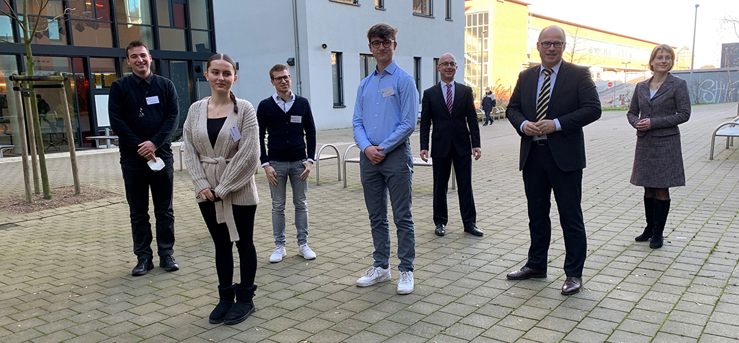 Das Bild zeigt sieben Personen vor einem modernen Gebäude. Die Personen haben alle ca. 1,5 Meter Abstand von einander.