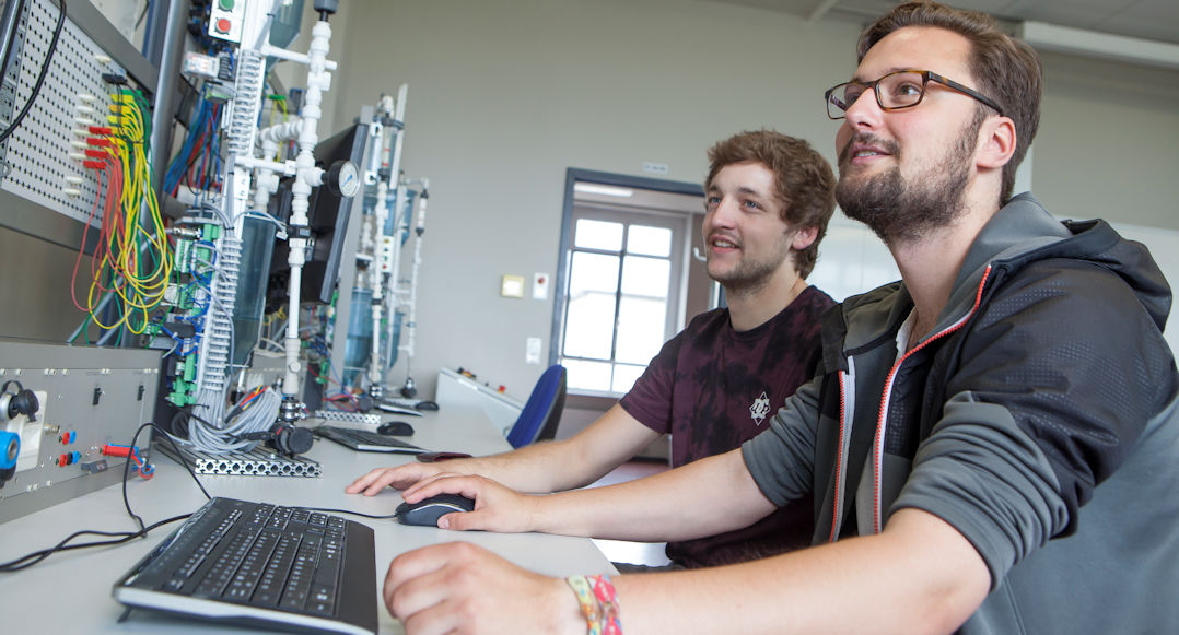 Das Bild zeigt zwei Personen die in einem Klassenraum nebeneinander vor einem Computer sitzen. Es sieht so aus, als seien die beiden in einem Klassenraum, der auf technischen Unterricht spezialisiert ist.