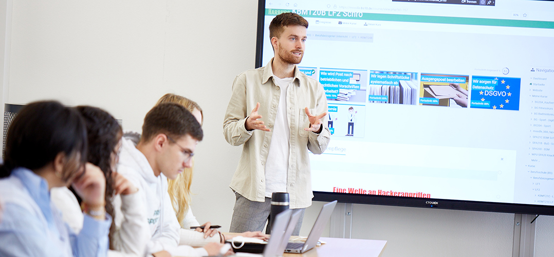 Auf dem Bild sind ingesamt fünf Personen zu sehen. Die vier Personen sitzen an der Seite an einem Tisch und haben einen Laptop vor sich stehen. Eine Person in der Mitte steht vor einem Bildschirm und gestikuliert, als ob er gerade eine Rede oder einen Vortrag hält. Der Raum ist hell.