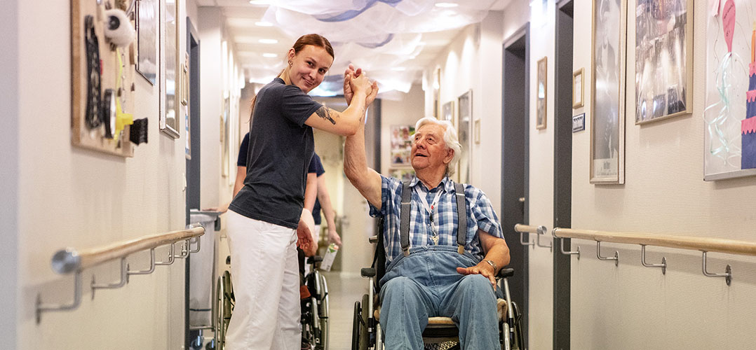 Auf dem Bild sind zwei Personen zu sehen, wovon eine Person im Rollstuhl sitzt und die andere neben ihr steht. Sie geben sich beide die Hand, so als ob sie einklatschen. Der Raum in dem sie sich befinden, scheint in einem Flur in einem Krankenhaus zu sein und ist künstlich beleuchtet.