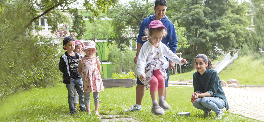 Auf dem Bild sind zwei Erwachsene und vier Kinder zu sehen, die draußen auf einer Wiese und bei Tageslicht scheinbar zusammen spielen. Im Hintergrund ist ein Schulgebäude und Bäume zu sehen.