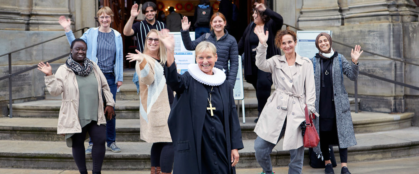 Auf dem Bild sind neun Personen auf einer Steintreppe und einer großen Tür zu sehen. Sie stehen draußen, vermutlich vor dem Eingang einer Kirche und winken in die Kamera. Es ist hell und in der Mitte steht eine Person die ein traditionelles Kirchenoutfit trägt und ein goldenes Kreuz und eine weiße Halskrause trägt.