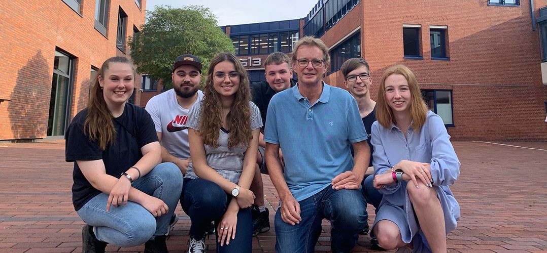 Auf dem Bild sind sieben Personen vor einem Schulgebäude zu sehen. Sie knien und schauen lächelnd in die Kamera. Sie tragen sommerliche Kleidung und im Hintergrund ist ein Baum zu sehen.