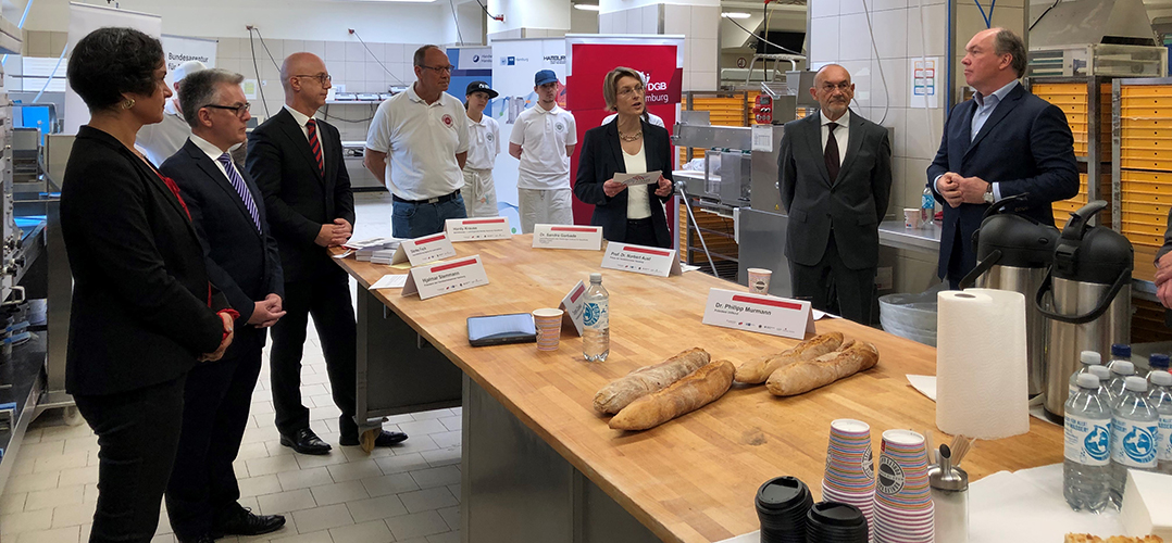 das Bild zeigt sieben Personen, die in einem Innenraum vor einem Tisch stehen, auf dem Namensschilder stehen und vier Brote sowie Wasserflaschen und Becher stehen. Im Hintergrund Werbebanner und zwei Personen in weißer Kleidung zusehen. Es sieht so aus, als sei ds Bild in einer Bäckerei aufgenommen worden.