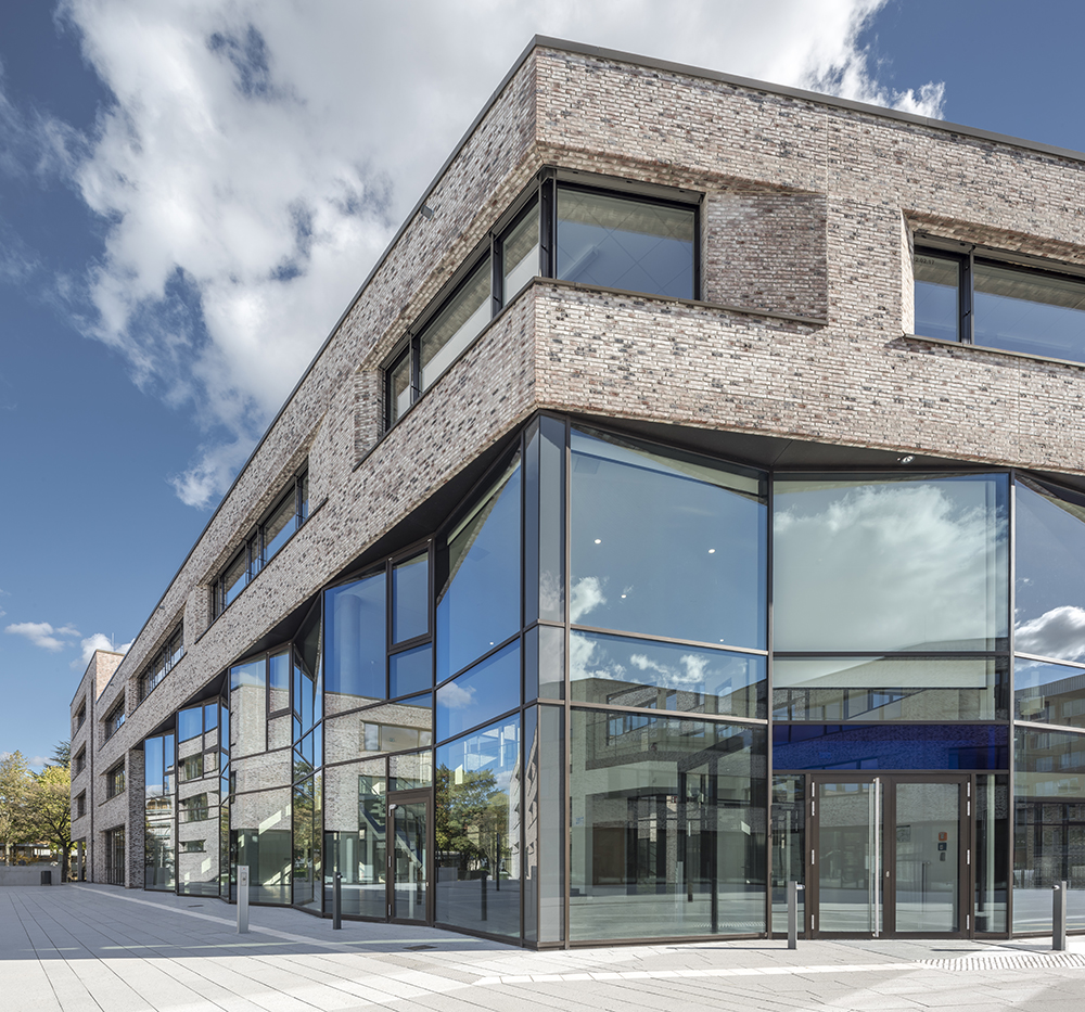 Das Bild zeigt einen modernen dreistöckigen Bau mit Flachdach, dessen untere zwei Stockwerke durch eine Reihe von schräg zueinander angeordneten Glasfronten besteht. So entsteht ein leichtes Zickzack-Muster. Das obere Stockwerk besteht aus braunem Klinker.