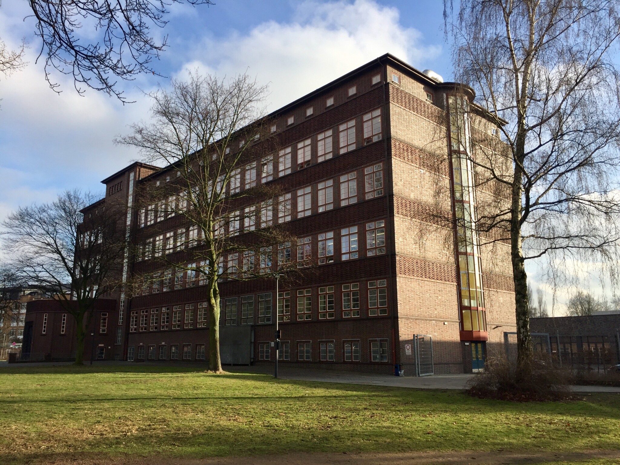 Das Bild zeigt einen fünfstöckigen Rotklinkerbau mit Flachdach und vielen Fenstern. Vor dem Gebäude befindet sich eine grüne Wiese mit einigen großen Bäumen.