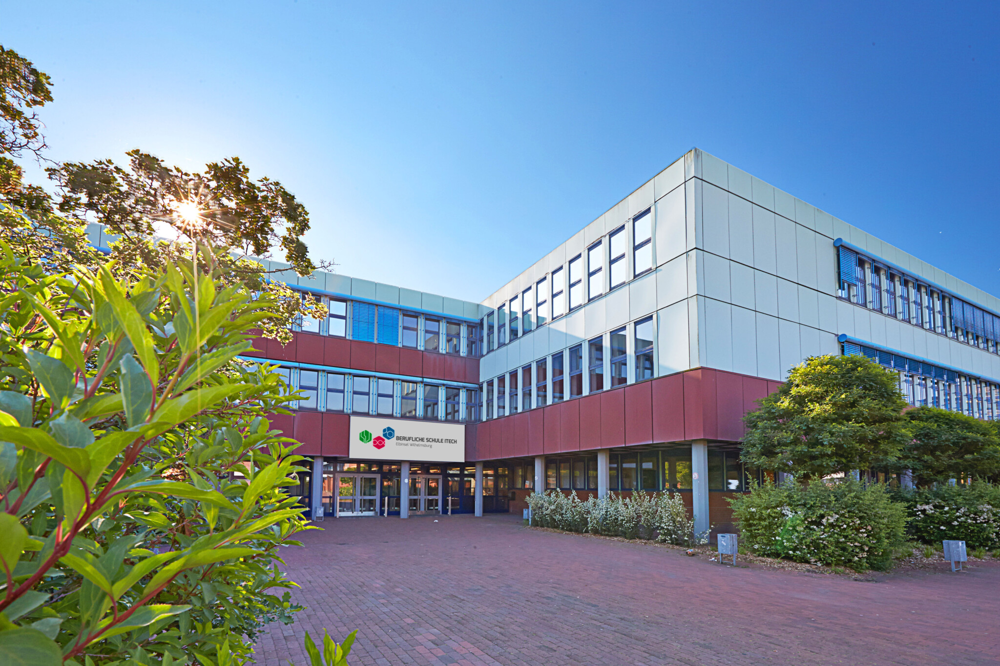 Das Bild zeigt due BS 14, einen dreistöckigen Schulbau in weiß und rot mit vielen Fenstern. Ein großes Schild mit dem Schulnamen markiert der Eingang. Der Schulhof ist rot gepflastert und mit Büschen und kleinen bäumen entlang des Gebäudes geschmückt. Einige grüne Blätter sind im Vordergrund am Bildrand zu sehen.