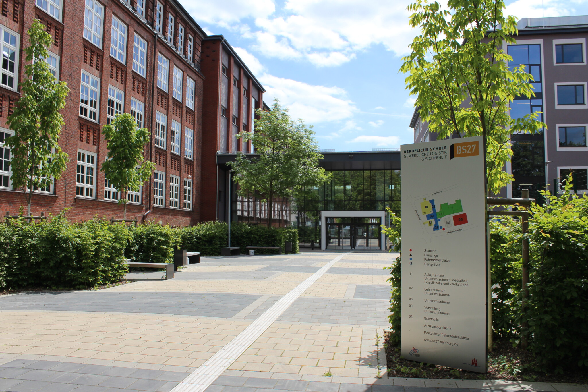 Zu sehen ist die BS27, die links aus einem Rotklinkerbau mit weißen Fensterrahmen und rechts aus einem braunen Neubau besteht. Die beiden Flügel werden durch einen gläsernen Eingangsbereich miteinander verbunden. Im Vordergrund ist ein gepflasterter Schulhof mit Büschen und Bäumen an den Rädern zu sehen. Ein großen Schild zeigt den Lageplan zur Orientierung auf dem Gelände.