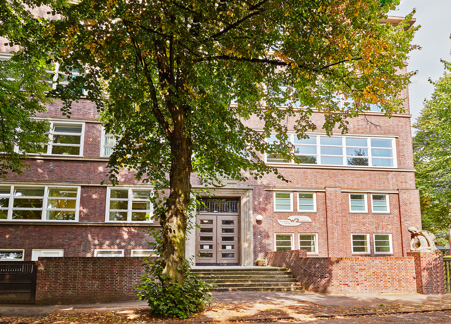 Das Bild zeigt im Vordergrund einen großen Baum mit dichten Blättern. Dahinter ist die BS29 zu sehen. Ein 4-stöckiger Rotklinker-Bau mit weißen Fensterrahmen und eine braunen Flügeltür. Eine kleine Backsteinmauer trennt das Schulgelände von der Straße.