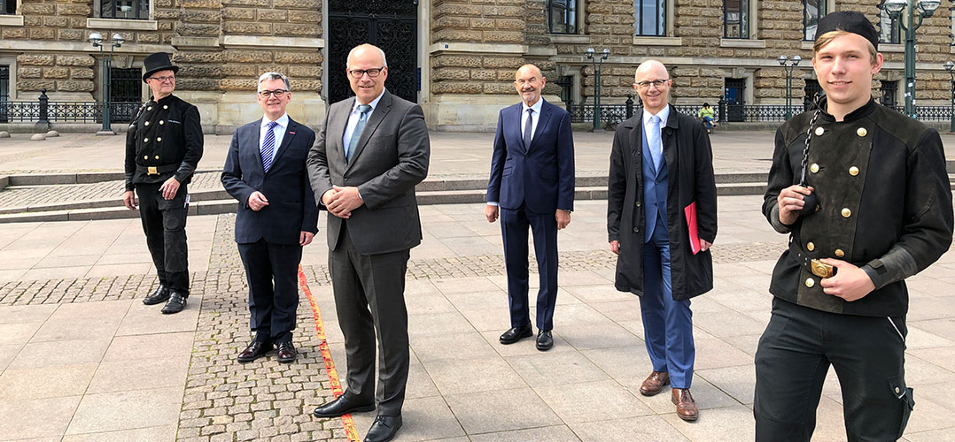 Das Bild zeigt sechs Personen, vor einem historischen Gebäude. Zwei der Personen tragen Schonsteinfegeruniformen, während die anderen in Anzügen gekleidet sind. Sechs Personen stehen auf einem gepflasterten Platz vor einem großen, steinernen Gebäude, dem Hamburger Rathaus, mit detaillierten architektonischen Merkmalen. Zwei der Personen,tragen Uniformen von Schornsteifegern. Vier Männer sind in formellen Anzügen gekleidet; ihre Kleidung variiert in Farbe und Stil. Der Himmel ist bewölkt, aber hell, was auf einen Tag mit bedecktem Himmel hindeutet.