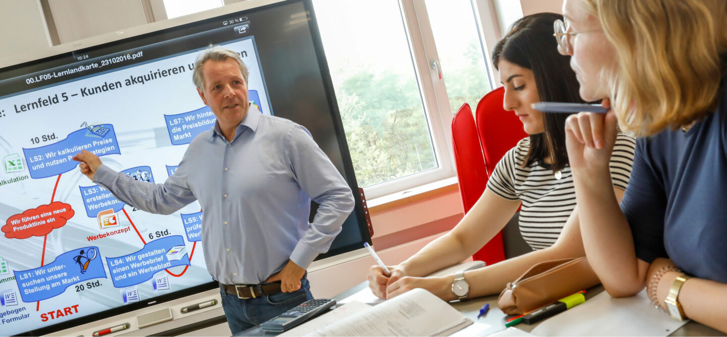 Das Bild zeigt eine Schulung oder einen Workshop in einem hellen Raum. Ein Trainer steht vor einem großen Bildschirm und erklärt den Inhalt, während zwei Teilnehmerinnen zuhören und Notizen machen. Der Trainer trägt ein hellblaues Hemd und zeigt mit einer Hand auf den Bildschirm. Zwei weibliche Teilnehmerinnen sitzen rechts im Bild. Auf dem Tisch vor den Teilnehmerinnen liegen Notizblöcke, Stifte und Marker. Der große Bildschirm zeigt eine Präsentation mit Text und Diagrammen in deutscher Sprache über Kundenakquise. Der Raum ist hell, mit großen Fenstern im Hintergrund, durch die Tageslicht einfällt. Es scheint, dass die Schulung sich auf Kundenakquise konzentriert, da der Bildschirm eine Präsentation mit Text und Diagrammen in deutscher Sprache darüber anzeigt. Die Notizblöcke und Stifte auf dem Tisch deuten darauf hin, dass die Teilnehmerinnen Notizen machen, um das Gelernte festzuhalten.