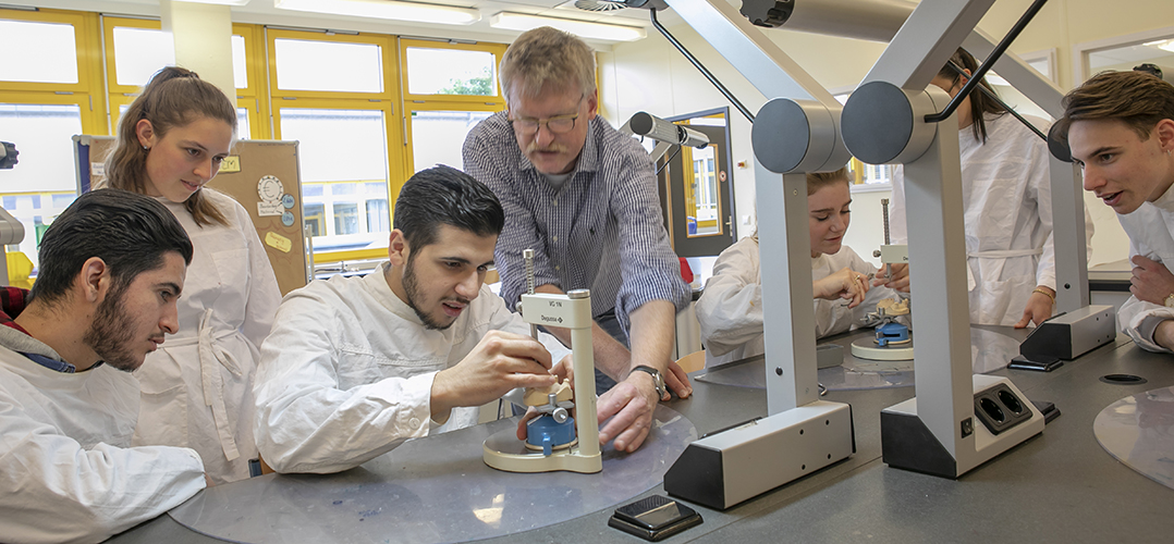 Das Bild zeigt eine Gruppe von Menschen, die in einem Labor oder Klassenraum arbeiten und Mikroskope benutzen. Das Labor bzw. der Klassenraumist gut beleuchtet und hat Fenster im Hintergrund. Die Personen tragen weiße Laborkittel und sind um einen Tisch gruppiert. Zwei Personen benutzen Mikroskope, die auf dem Tisch stehen. Es gibt ein komplexes Gestell oder Gerät mit mehreren Armen und Verbindungen über dem Tisch; seine Funktion ist nicht klar ersichtlich. Im Hintergrund sind Fenster sichtbar, durch die Tageslicht einfällt.