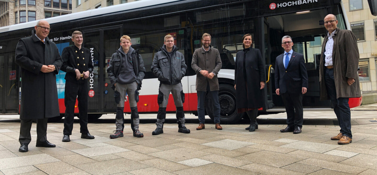 Das Bild zeigt sieben Personen, vor einem Bus mit der Aufschrift “HOCHBAHN”. Die Personen tragen verschiedene Kleidungsstile, darunter Anzüge, Freizeitkleidung und eine Schornsteinfegeruniform. Im Hintergrund ist ein Bus mit der Aufschrift “HOCHBAHN” und “für Hamburg” zu sehen. Der Himmel erscheint bewölkt und das Umgebungslicht ist diffus. Gebäude sind im Hintergrund sichtbar, aber nicht klar erkennbar wegen des Busses und der Personen im Vordergrund.