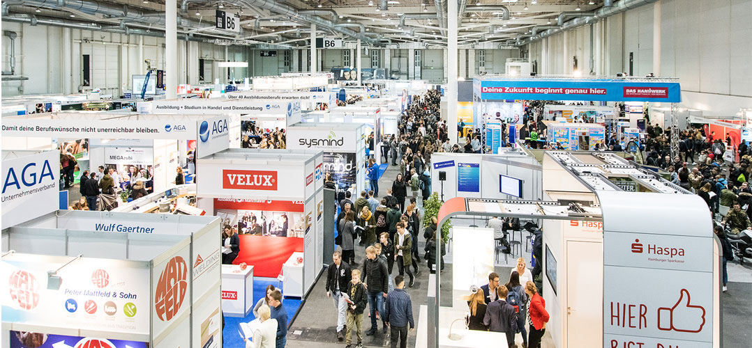 Das Bild zeigt eine belebte Messehalle mit verschiedenen Ausstellungsständen und einer Menschenmenge. Die Messehalle ist groß und geräumig, mit hohen Decken und Beleuchtung, die den gesamten Raum erhellt. Es gibt viele Ausstellungsstände mit Bannern und Logos verschiedener Unternehmen wie AGA, sysmind, VELUX und Haspa. Die Stände sind gut organisiert und bieten Informationen über die ausgestellten Produkte oder Dienstleistungen. Eine große Anzahl von Menschen ist zu sehen; sie bewegen sich durch die Gänge oder stehen an den Ständen, um sich zu informieren oder zu interagieren. Über den Ständen hängen Schilder mit Aufschriften wie “B6” zur Orientierung innerhalb der Halle. Ein großes Banner im Hintergrund trägt die Aufschrift “Deine Zukunft beginne genau hier”