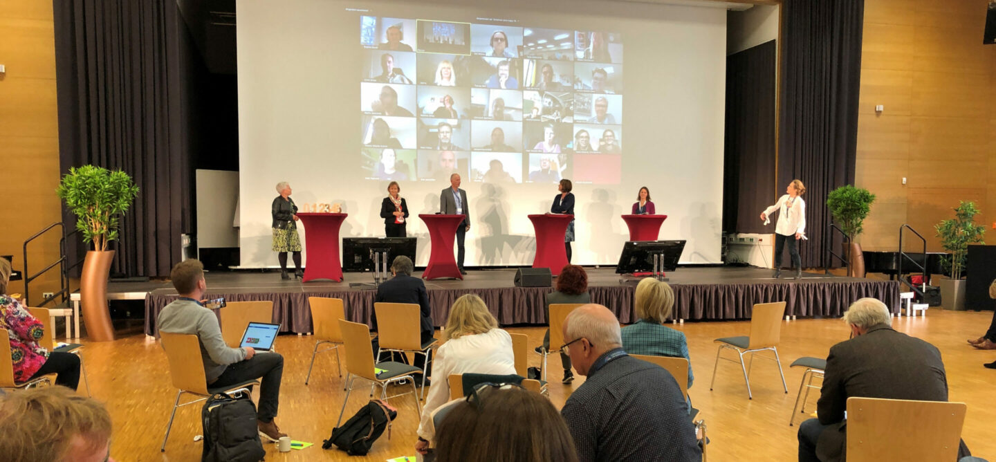 Das Bild zeigt eine Konferenz oder Veranstaltung in einem großen Saal. Auf der Bühne stehen fünf Personen, und über ihnen wird eine Live-Übertragung von verschiedenen Teilnehmern auf einer großen Leinwand angezeigt. Der Raum ist groß und geräumig mit einem Holzboden und einer hohen Decke. Es gibt viele Stühle, auf denen die Teilnehmer sitzen; einige sind besetzt, andere leer. Fünf Personen stehen auf der Bühne; jede steht hinter einem separaten Rednerpult. Eine große Leinwand zeigt ein Zoom-Meeting oder eine ähnliche Video-Konferenz mit vielen Teilnehmern in einzelnen Fenstern. Es gibt Pflanzen zu beiden Seiten der Bühne, die zur Dekoration dienen. Einige Teilnehmer haben Laptops und machen sich Notizen oder arbeiten während der Veranstaltung