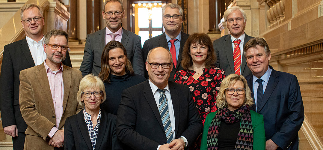 Auf dem Bild sind mehrere Personen zu sehen. Sie tragen formelle Kleidung und stehen in einem Gebäude mit einer Treppe im Hintergrund. Die Personen tragen verschiedene Arten von Kleidung, darunter Anzüge, Blusen und Kleider. Das Ambiente wirkt elegant und formell, was durch die Beleuchtung und die Struktur des Gebäudes hervorgehoben wird.