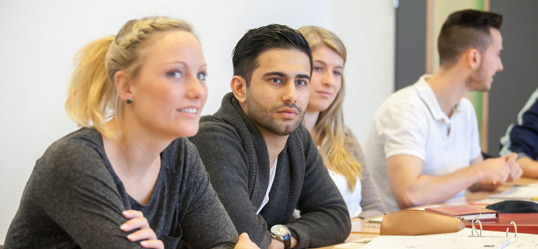 Das Bild zeigt mehrere Personen, die an einem langen Tisch sitzen und sich auf Papierdokumente konzentrieren. Die Personen scheinen in einem Schulunterricht zu sein, da sie Dokumente vor sich haben. Es gibt verschiedene Gegenstände auf dem Tisch, darunter Papiere und ein Ringbuch. Die Kleidung der Personen ist lässig; sie tragen keine formelle Kleidung oder Uniformen. Der Hintergrund des Bildes ist einfach und unauffällig, was den Fokus auf die am Tisch sitzenden Personen lenkt.