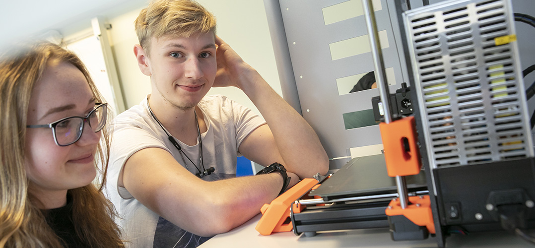 Das Bild zeigt zwei Personen, die einen 3D-Drucker betrachten. Der Raum, in dem sie sich befinden, ist hell beleuchtet und hat blaue Wände. Der 3D-Drucker ist groß und modern, mit einer grauen Struktur und orangefarbenen Akzenten. Es sieht so aus, als ob die Personen den Druckvorgang beobachten oder darüber diskutieren.