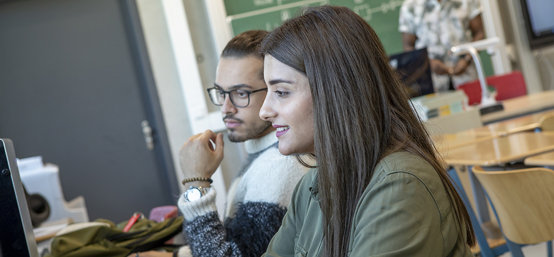 Das Bild zeigt zwei Personen, die in einem Klassenzimmer vor Computern sitzen. Eine Person trägt eine grüne Jacke und hat langes Haar. Im Hintergrund sind ein schwarzes Whiteboard und eine Tür sichtbar.