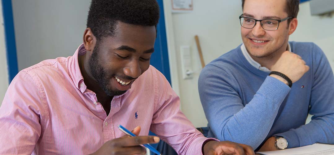 Das Bild zeigt zwei Personen, die an einem Tisch sitzen und sich auf ein Dokument konzentrieren. Die Person links trägt ein rosa gestreiftes Hemd und hält einen blauen Stift. Die Person rechts ist in einem blauen Pullover gekleidet und trägt eine Uhr am linken Handgelenk. Beide schauen auf ein Dokument oder Buch, das auf dem Tisch liegt, aber der Inhalt ist nicht sichtbar. Im Hintergrund ist eine weiße Wand mit einer blauen Tür oder einem Fenster zu sehen.