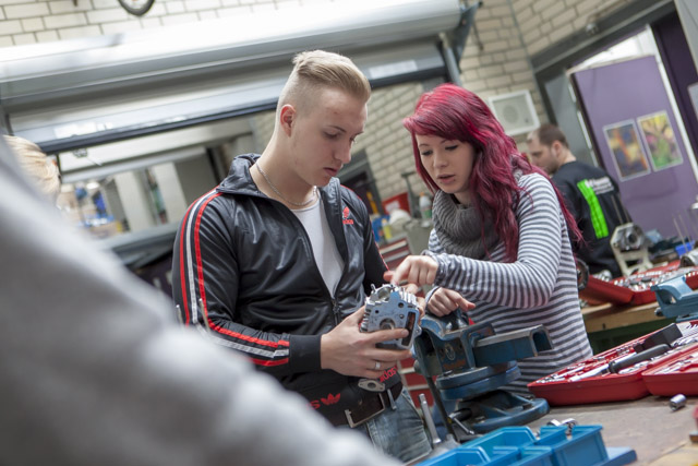 Das Foto zeigt einen jungen blonden Mann und eine junge rothaarige Frau, die an einer werkbank stehen. Der Mann hat ein Gerät aus Metall in der Hand, auf das die Frau zeigt.