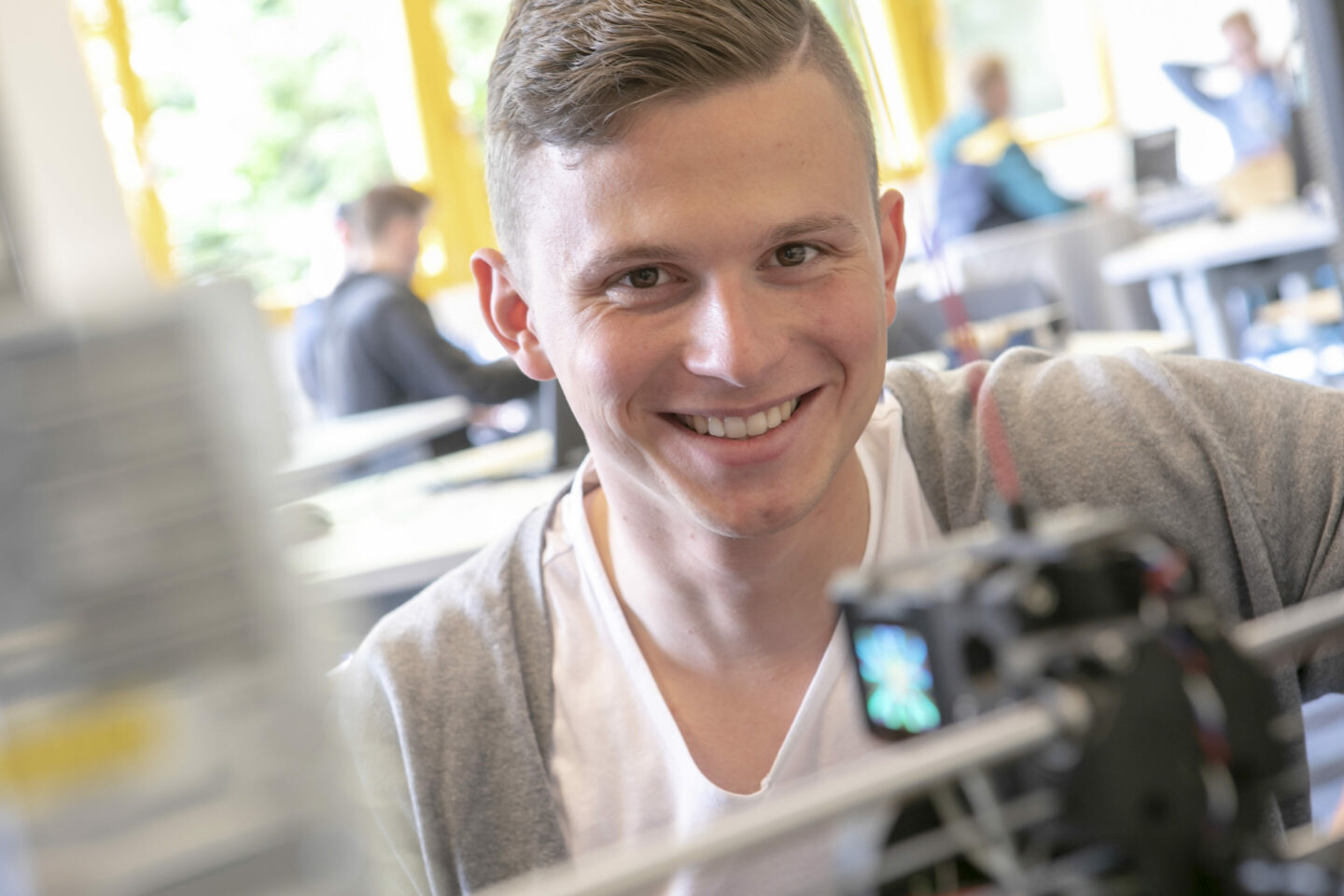 Das Bild zeigt eine junge männliche Person, in einem hellen Raum mit anderen Menschen im Hintergrund. Die Person scheint sich auf eine Maschine oder ein Gerät zu konzentrieren. Es gibt Fenster mit gelben Vorhängen, durch die viel natürliches Licht einfällt. Ein Teil einer Maschine oder eines Geräts ist im Vordergrund sichtbar; es könnte sich um eine Art von technischem oder wissenschaftlichem Equipment handeln.