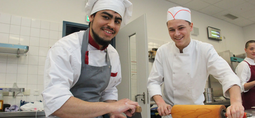 Das Bild zeigt zwei Köche in einer professionellen Küche, die Essen zubereiten. Der Koch auf der linken Seite trägt eine traditionelle weiße Kochjacke mit roten Akzenten und eine hohe weiße Kochmütze. Der Koch auf der rechten Seite trägt eine vollständig weiße Uniform mit einer niedrigeren Mütze, die mit einem roten Band verziert ist. Beide Köche arbeiten an einem Tisch; der linke schält Gemüse und der rechte rollt Teig aus. Im Hintergrund sind Küchengeräte und Utensilien sichtbar, darunter ein Edelstahlspülbecken und Regale mit verschiedenen Gegenständen.