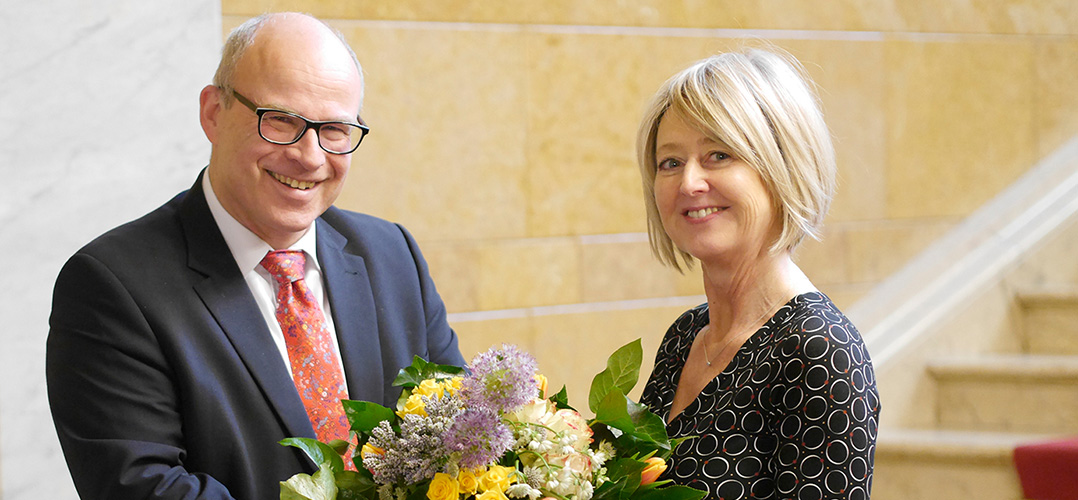 Das Bild zeigt zwei Personen in einem eleganten Innenraum. Die Person auf der linken Seite trägt einen dunklen Anzug mit einer gemusterten Krawatte und hält einen bunten Blumenstrauß. Die Person auf der rechten Seite ist in ein Kleid mit einem kreisförmigen Muster gekleidet. Der Hintergrund zeigt eine Treppe und eine Wand aus gelbem Marmor oder Stein, was auf eine elegante Umgebung hinweist.