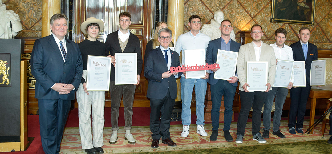 Das Bild zeigt eine Gruppe von Menschen, die Auszeichnungen in den Händen halten. Sie stehen in einem prächtig dekorierten Raum im Hamburger Rathaus. Jede Person hält ein Zertifikat oder ein Schild. Die Personen tragen formelle Kleidung, darunter Anzüge und Kleider. Der Raum ist luxuriös gestaltet mit aufwendigen Wandverzierungen, Gemälden und einer Statue im Hintergrund. Ein roter Teppich bedeckt den Boden, und ein prunkvoller Kronleuchter ist teilweise sichtbar. Auf einem der Schilder steht “#wirfeiernhandwerk