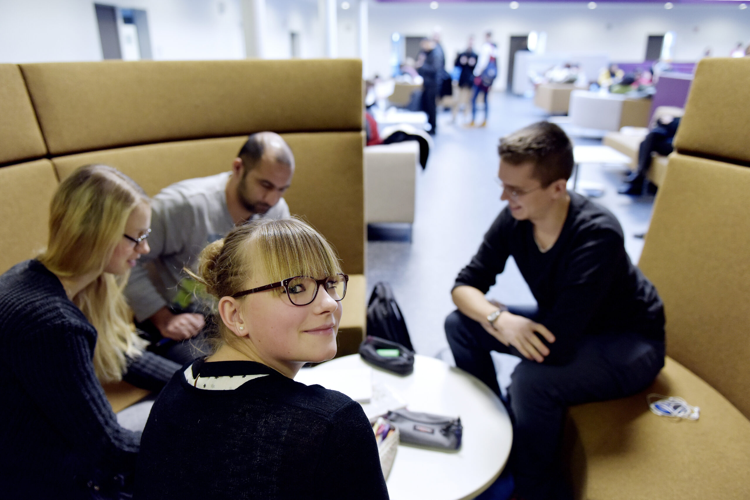 Das Bild zeigt 4 junge Menschen, die in einer kreisrunden beigefarbenen Sitzecke um einen runden Tisch sitzen, die hinteren beiden Männer und eine Frau sind leicht verschwommen. Im Vordergrund schaut eine blonde Frau mit Brille einen über ihre Schulter zurückschauend an.