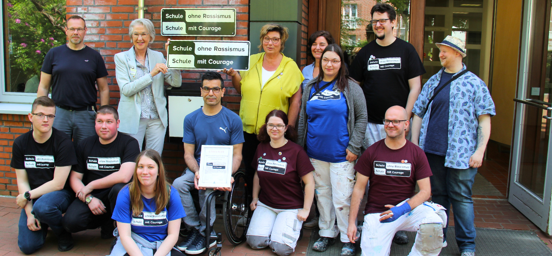 Das Bild zeigt eine Gruppe von Menschen, die vor einem Backsteingebäude stehen. Einige Personen tragen T-Shirts mit der Aufschrift “Schule ohne Rassismus - Schule mit Courage”. Eine Person hält ein Schild hoch, auf dem ebenfalls “Schule ohne Rassismus - Schule mit Courage” steht. Eine andere Person hält eine Urkunde oder ein Zertifikat in den Händen. Die Personen stehen auf einem gepflasterten Weg vor dem Eingang des Gebäudes.