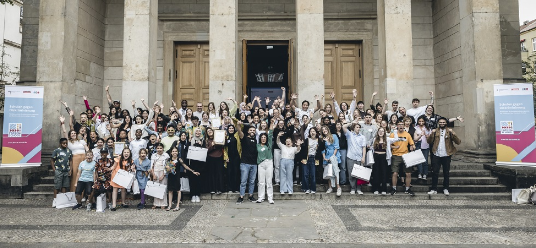 Das Bild zeigt eine große Gruppe von Menschen, die vor einem Gebäude mit Säulen und Stufen stehen. Sie scheinen fröhlich und aufgeregt zu sein, viele heben ihre Hände oder Arme. Es gibt auch Banner an den Seiten des Eingangs. Das Gebäude hat ein klassisches Design mit hohen Säulen, die den Eingangsbereich rahmen. Der Himmel ist bewölkt, aber hell. Es handelt sich um eine Veranstaltung einer Schule und die Menschenmenge könnte für eine Feier, eine Zeremonie oder einen besonderen Anlass versammelt sein.