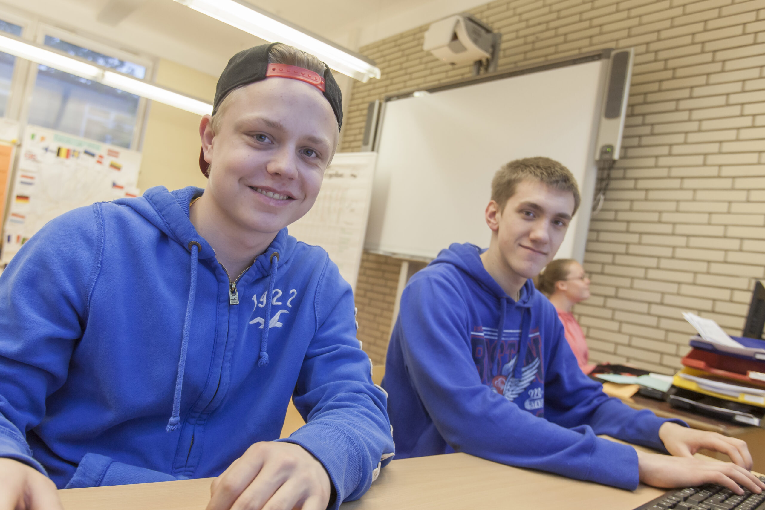 Das Bild zeigt 2 männliche Jugendliche an einem Schultisch sitzen. Der rechts sitzende jugendliche bedient eine Tastatur. Beide haben ein blaues Sweat-Shirt an. Der links sitzende Jugendliche trägt eine Baseball-Kappe verkehrt herum.