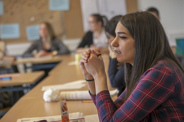 Das Bild zeigt eine an Tischen sitzende Schulklasse. Die hinteren Schülerinnen und Schüler sind verschwommen. Im Vordergrund sitzt eine junge dunkelhaarige Frau an einem Tisch, schaut nach links und stützt ihre Ellenbogen auf den Tisch.