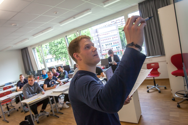Das Bild zeigt im Hintergrund einen Klassenraum, in dem 6 junge Männer an Schultischen sitzen. Im Vordergrund steht ein junger Mann mit blauem Oberteil, der einen Stift in der Hand hält und etwas an eine Tafel zu schreiben scheint. Die Tafel ist nicht zu sehen.