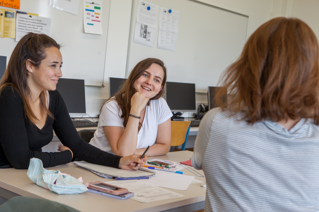 Das Bild zeigt 3 junge Frauen, die um einen Schultisch sitzen, auf dem Zettel und Stifte liegen. von der vorderen braunhaarige Frau sieht man nur den Rücken. Die beiden anderen dunkelhaarigen Frauen, eine mit schwarzem und eine mit weißem Oberteil lächeln diese an.
