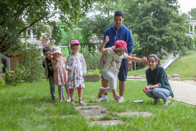 Das Bild zeigt 4 Kinder im Vorschulalter, die auf einem Rasen hintereinander stehen. Es gibt im Rasen 5 Bodenplatten, Das vordere Kind springt von einer Platte zu einer anderen. Hinter den Kindern stehen 2 junge Erwachsene, ein stehender Mann und eine hockende Frau , die lächelnd auf die Kinder aufpassen.
