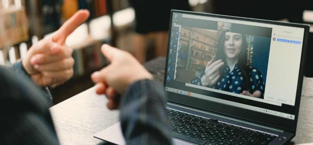 Das Bild zeigt zwei Personen, die vor einem Laptop sitzen. Der Laptop steht auf einem Tisch und zeigt einen aktiven Videoanruf mit einer anderen Person. Auf dem Bildschirm ist ein Videoanruf zu sehen. Die beiden Personen sind nicht direkt erkennbar, aber ihre Hände zeigen gestikulierend. Die Person im Videoanruf trägt ein gepunktetes Oberteil. Im Hintergrund des Videoanrufs sind Bücherregale erkennbar. Der Raum, in dem sich die beiden Personen befinden, ist unscharf und daher nicht detailliert erkennbar.