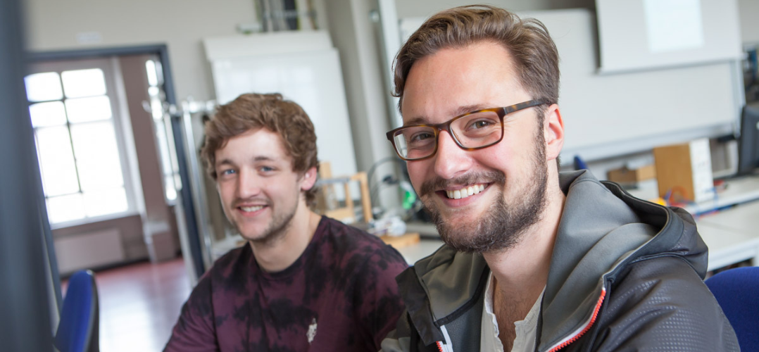 Zwei junge Männer lächeln freundlich in die Kamera. Sie sind bis zur Brust zu sehen. Beide haben braune Haare und helle Haut, der Mann im Vordergrund trägt eine Brille und einen gestutzten Vollbart. Im Hintergrund erahnt man einen Unterrichtsraum.