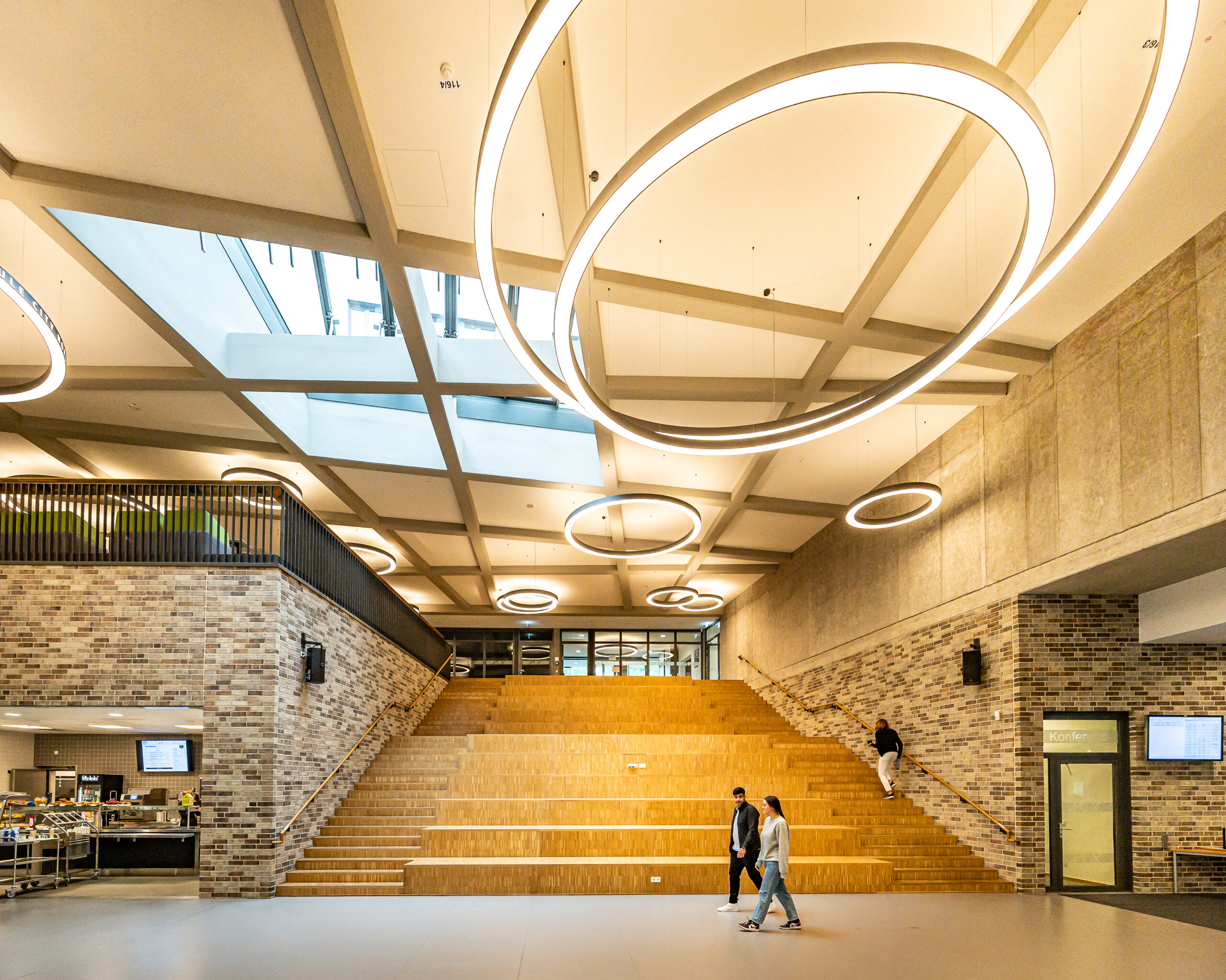 Ein heller und moderner Eingangsbereich mit einer Holztreppe, Betonwänden und sehr hohen Decken. Es hängen Kreisrunde Lampen unter der Decke und ein großes Oberlicht aus Glas lässt den Raum hell wirken. Vereinzelt sind Menschen zu sehen.