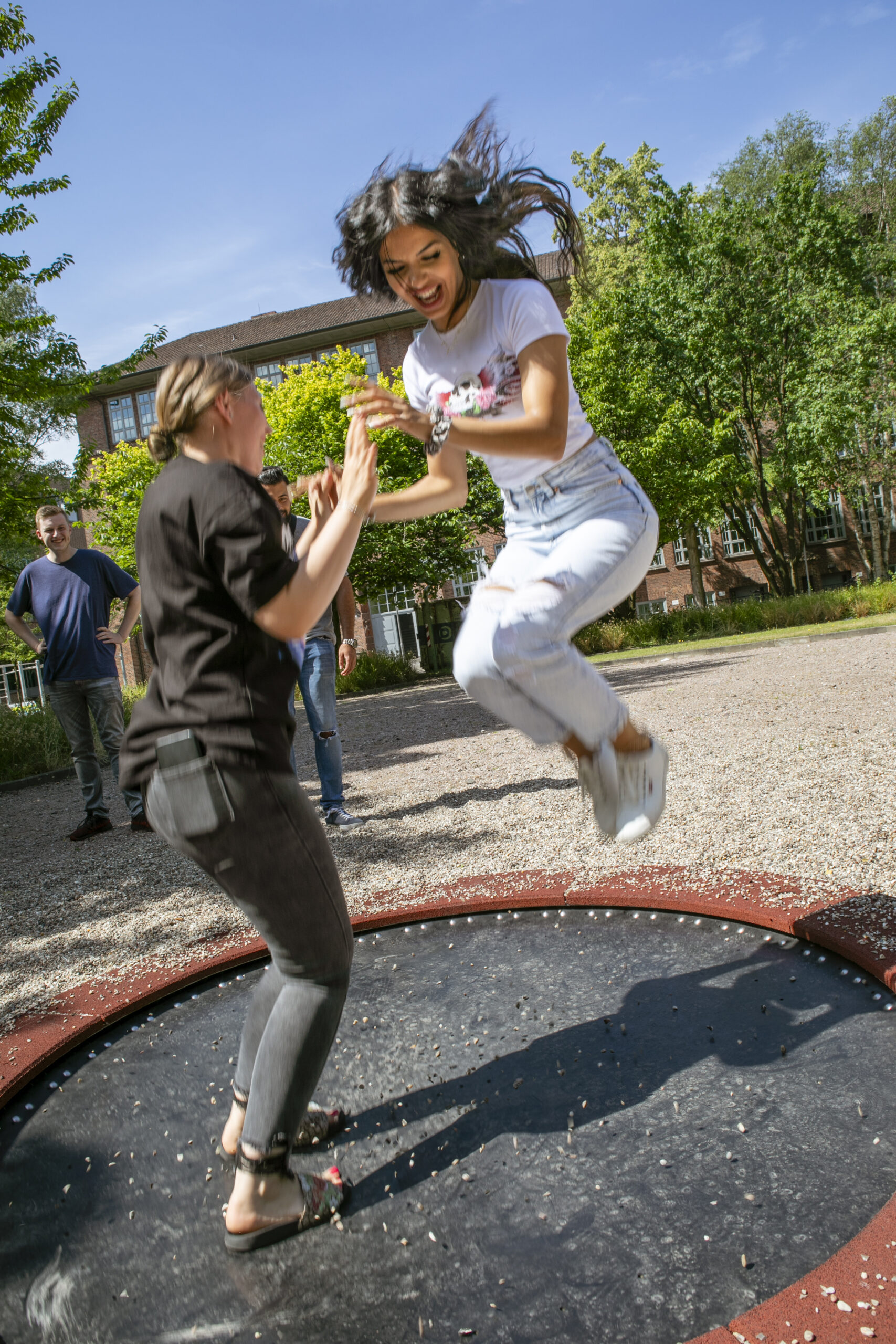 Das Foto zeigt 2 junge Frauen auf einem Trampolin auf einem Schulhof. Die rechte junge Frau ist weiß gekleidet und ist angewinkelten Beiden springend in der Luft, die linke Frau ist dunkel gekleidet, steht noch auf dem Trampolin und hält die Hände der anderen jungen Frau.